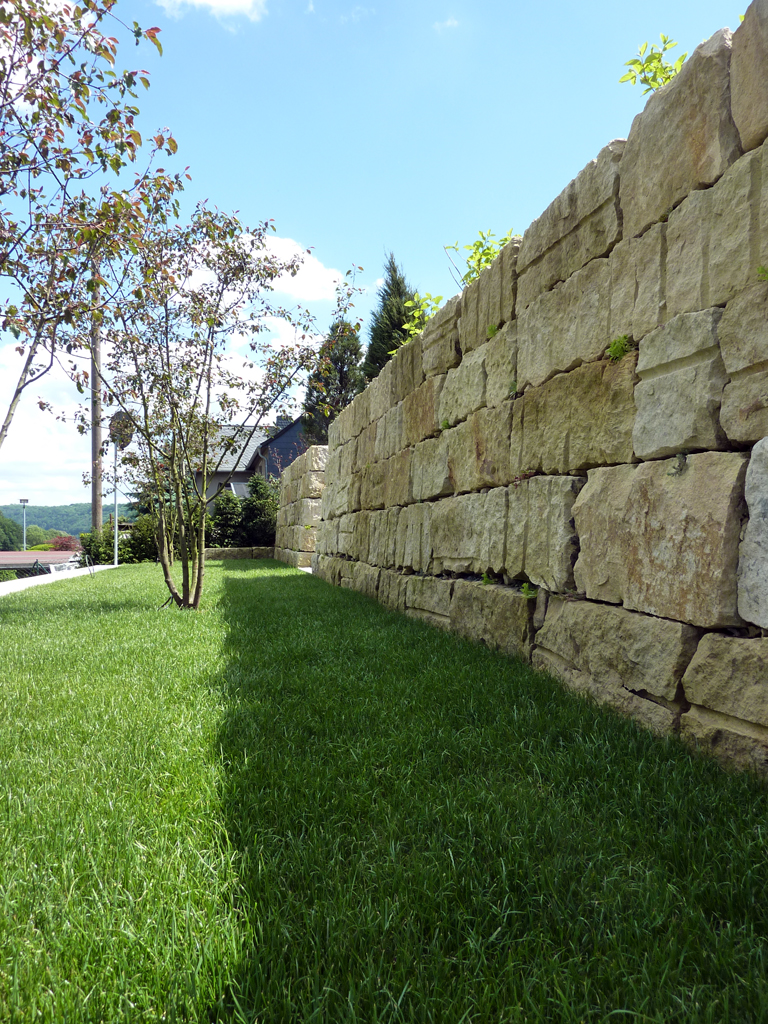 Eine steinerne Stützmauer verläuft entlang eines grünen Rasens mit einem kleinen Baum, unter einem strahlend blauen Himmel mit vereinzelten Wolken. Im Hintergrund sind einige Sträucher und ein Haus zu sehen.