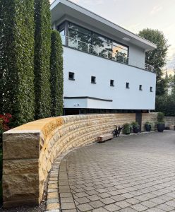 A modern two-story house with large windows, a light blue and beige exterior, a curved stone wall, potted plants, and a paved driveway, surrounded by tall trees and green spaces.