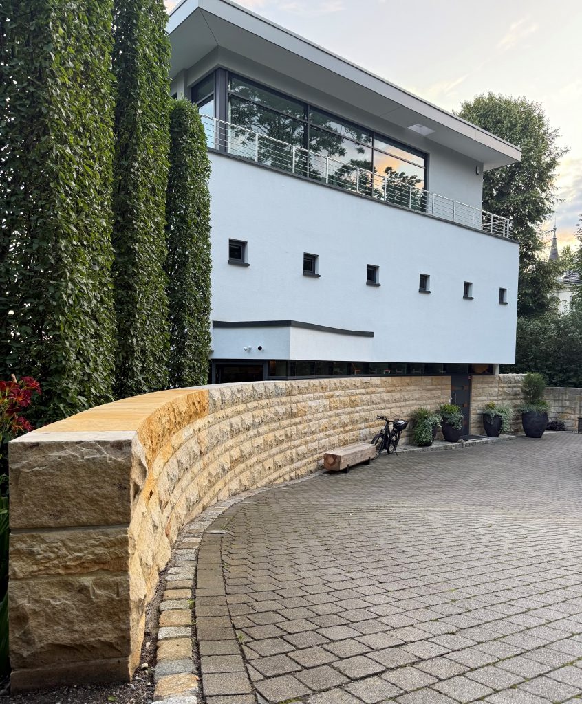 Modernes zweistöckiges Haus mit großen Fenstern, hellblauem und beigem Äußeren, geschwungener Steinmauer, Topfpflanzen und einer gepflasterten Auffahrt, umgeben von hohen Bäumen und Grünanlagen.