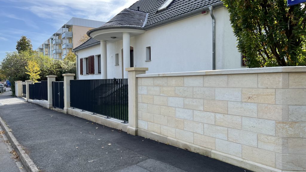 Ein modernes weißes Haus mit einem dunklen Ziegeldach und Säulen steht hinter einer beigen Steinmauer mit einem schwarzen Metalltor an einer ruhigen Wohnstraße. Im Hintergrund sind Bäume und angrenzende Gebäude zu sehen.