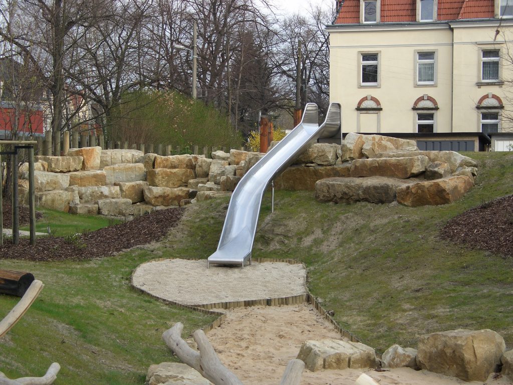 Eine Spielplatzrutsche aus Metall führt einen Grashügel hinunter, beginnt auf einer in große Felsen gebauten Plattform und endet in einer Sandfläche. Im Hintergrund sind blattlose Bäume und ein gelbes Gebäude zu sehen.
