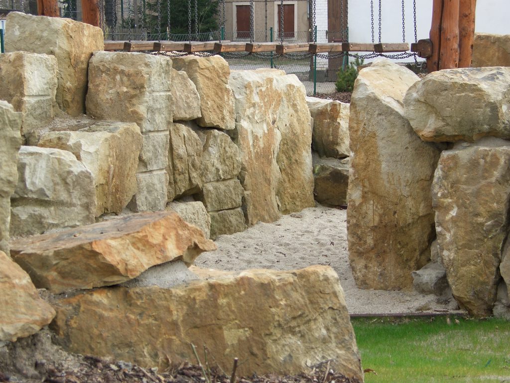Große, gestapelte Steinblöcke bilden einen Durchgang mit sandigem Boden, umgeben von Gras und Holzstrukturen im Hintergrund.