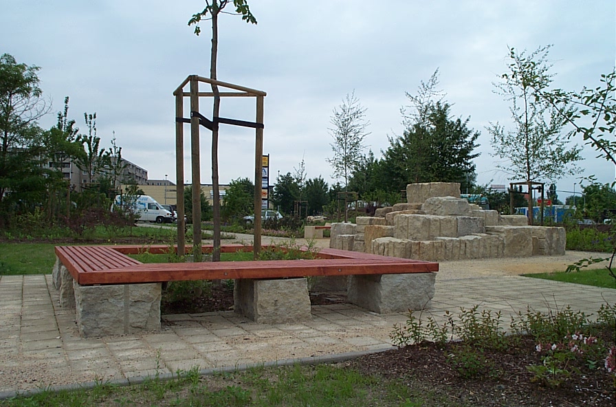 Ein kleiner Park mit einer Holzbank, die einen jungen Baum umgibt, Steinbänken und einer pyramidenförmigen Kletterstruktur aus Stein. Es gibt Bäume, Büsche und einen Weg, mit einem Lieferwagen und Gebäuden im Hintergrund.