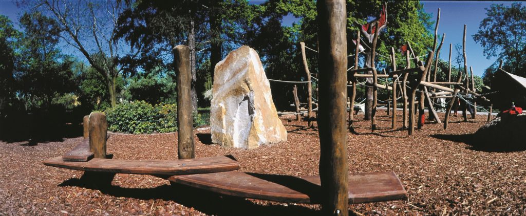 Ein hölzerner Spielplatz mit Klettergerüsten und einer Wippe ist von Bäumen und Mulch umgeben und steht unter einem strahlend blauen Himmel.