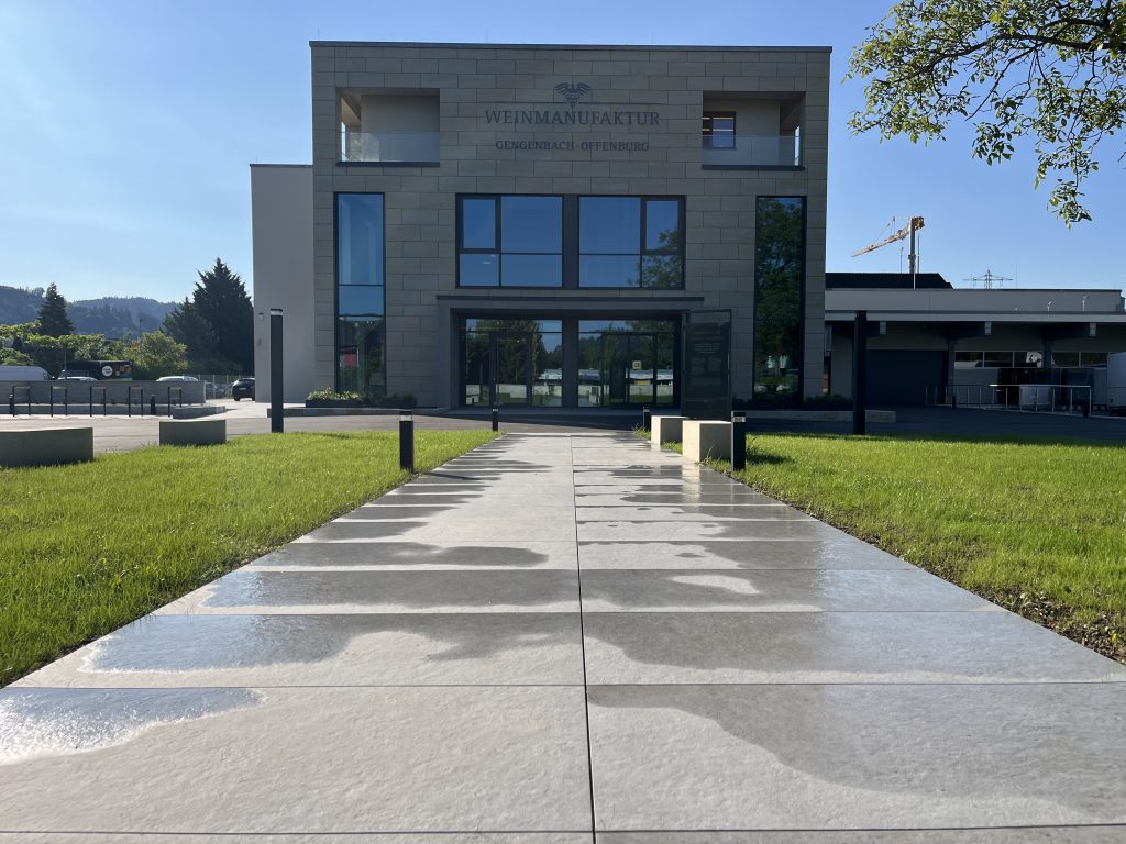 Ein modernes Gebäude mit großen Glasfenstern und der Aufschrift "Weinmanufaktur" steht am Ende eines nassen, sonnenbeschienenen Weges, umgeben von grünem Gras und ein paar Bäumen unter einem klaren blauen Himmel.