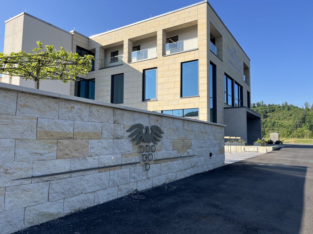 Ein modernes, beigefarbenes Steingebäude mit großen Fenstern und Balkonen steht hinter einer niedrigen Steinmauer mit einem metallenen Caduceus-Symbol, das auf eine medizinische Einrichtung hinweist. Der Himmel ist klar, und das Gelände ist von Grünflächen umgeben.