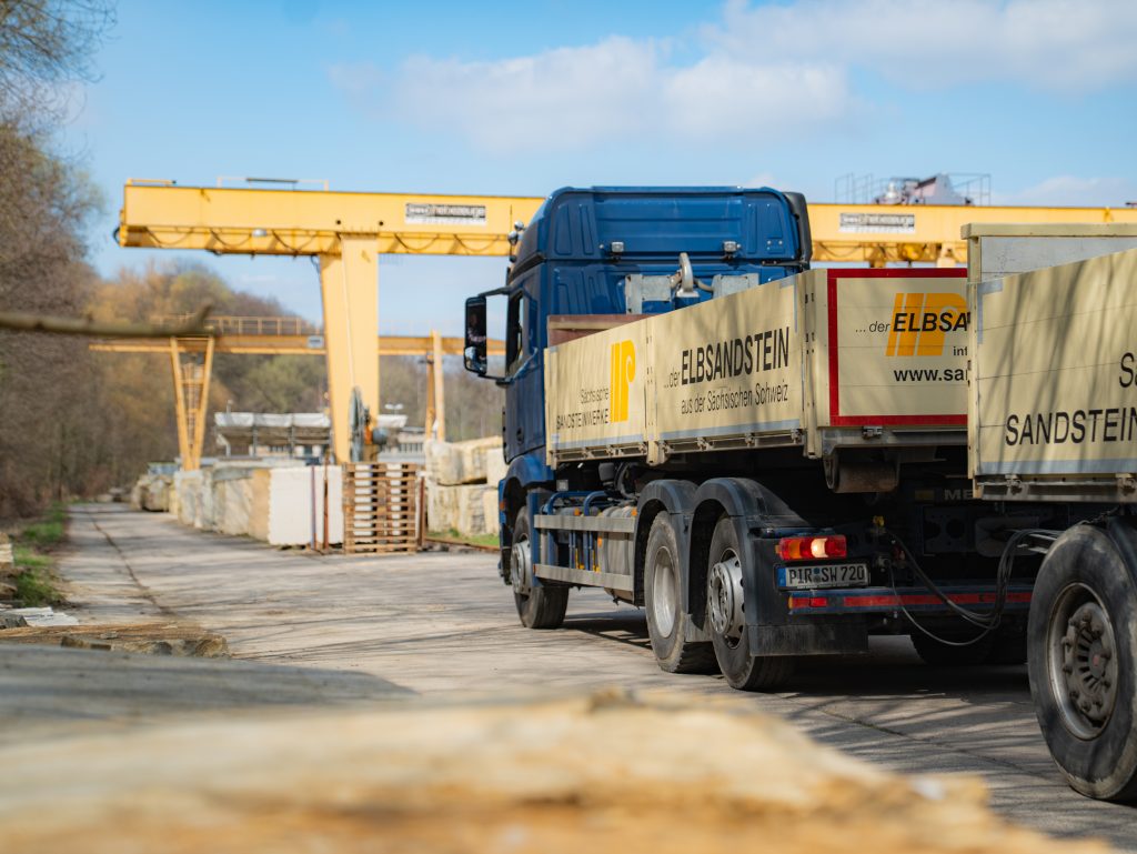 A blue truck, loaded with large blocks of stone, is parked on a paved road in an industrial park; yellow cranes and stacks of stone slabs can be seen in the background, and a blue sky stretches out above it all.