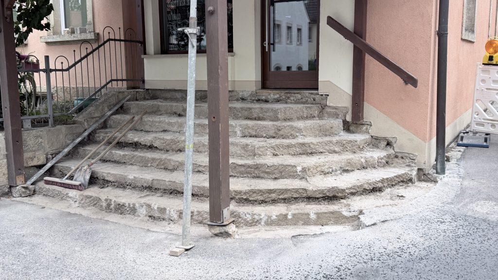 Abgenutzte Betonstufen, die gerade renoviert werden, mit Baugeräten und einer Metallstange daneben, vor dem Eingang eines pfirsichfarbenen Gebäudes.