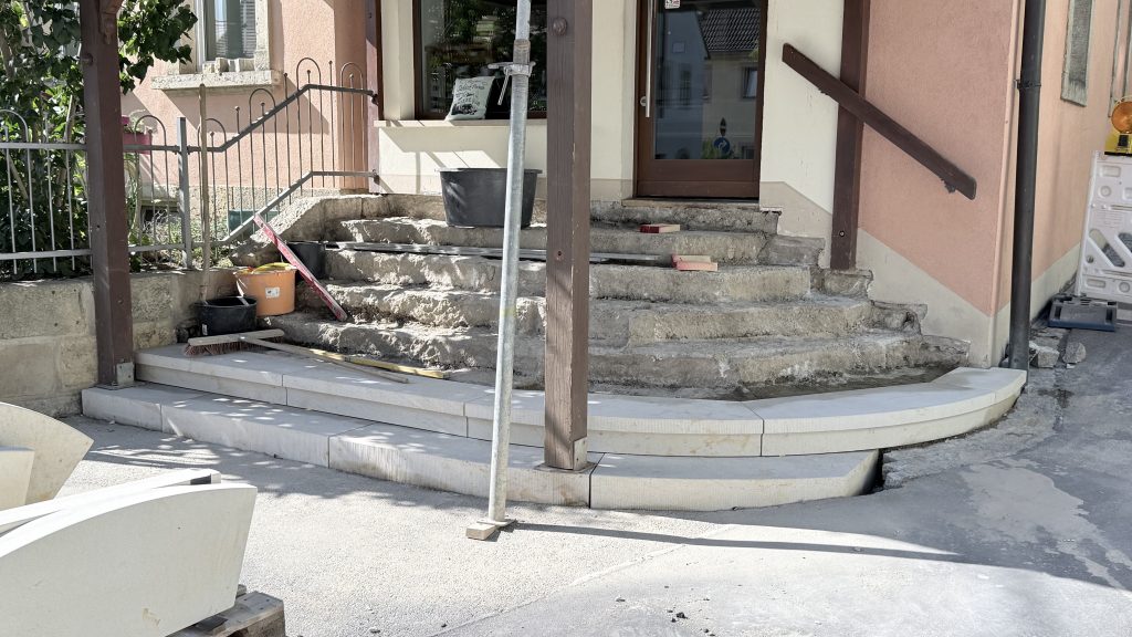 Ein Gebäudeeingang mit abgenutzten Steinstufen, der gerade renoviert wird. Am Sockel werden neue Steinplatten verlegt, und in der Nähe liegen Bauwerkzeuge und Baumaterialien verstreut. Davor stehen ein Holzpfosten und ein Metallgerüst.