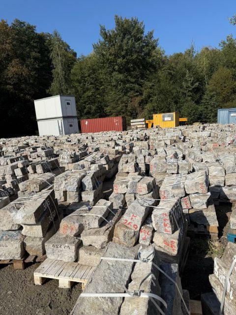 Auf einem Lagerplatz im Freien sind zahlreiche Holzpaletten gestapelt, auf denen graue Granitblöcke liegen, die mit roten Zahlen gekennzeichnet sind.
