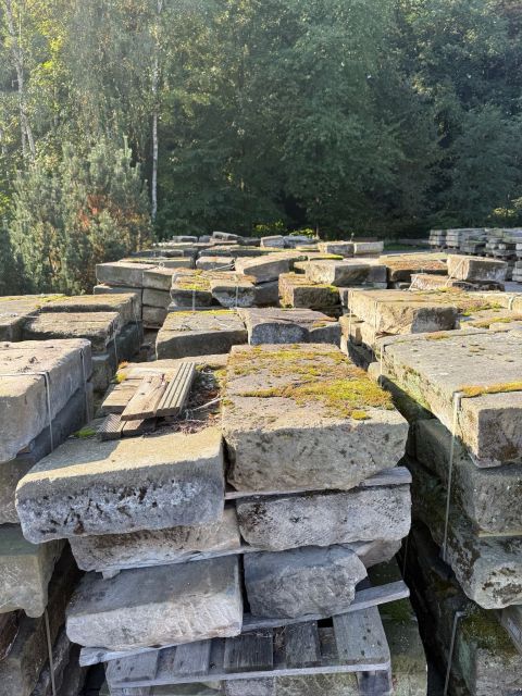 Große, moosbewachsene Steinblöcke, die in einem Waldgebiet auf Holzpaletten gestapelt sind.
